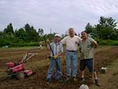 Three men pose for the camera in the middle of a freshly-tilled garden.