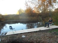 A man smokes sitting on a wooden dock overlooking a small lake.